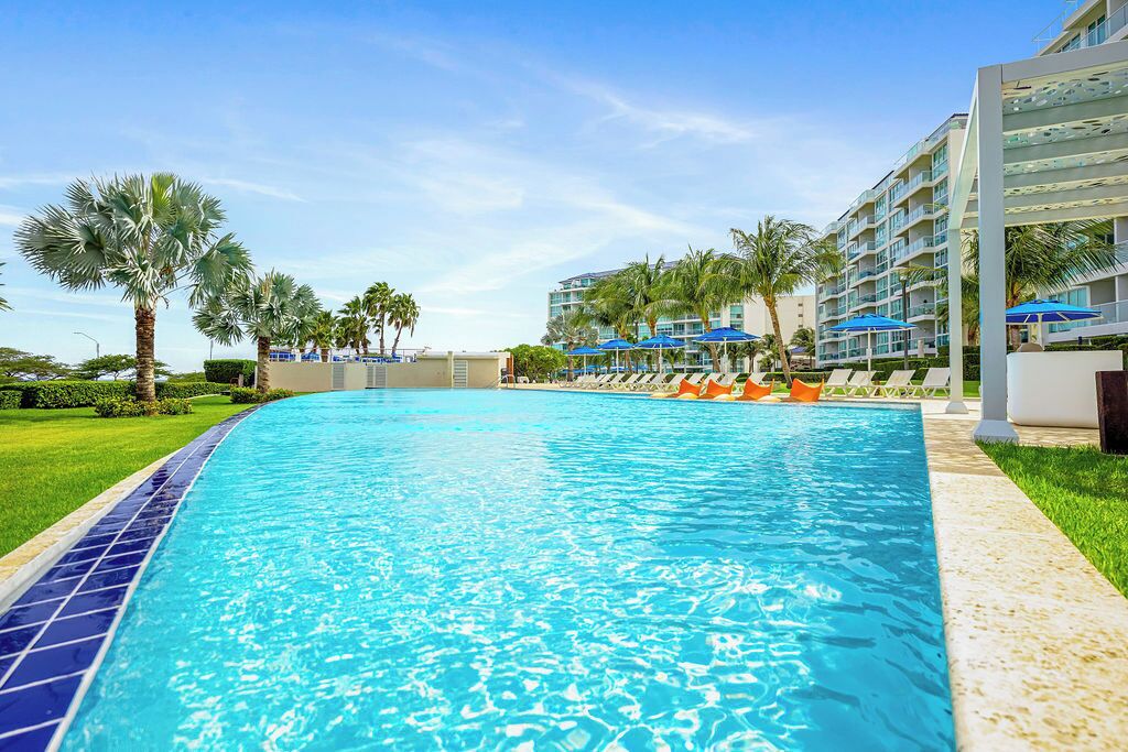 A swimming pool with lounge chairs and umbrellas, surrounded by palm trees and a hotel building.
