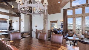 A spacious dining area with a wooden table and leather chairs, a chandelier, and large windows offering a view of the outdoors. - Luxury Big Sky Mountain Home With Private Lodge, Pool & Ski in/out access (Big Sky)