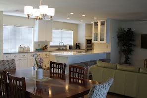 A modern kitchen with white cabinets, a dining area with a wooden table and chairs, and a living area with a sofa and a potted plant.