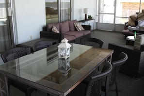 A glass-top dining table with wicker chairs, a cushioned sofa, and a lamp in a bright room with large windows.