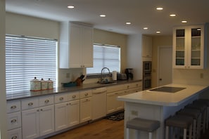 A modern kitchen with white cabinetry, a central island with stools, a built-in oven, and a window with blinds.