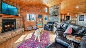 Interior acogedor de una cabaña con chimenea de piedra, mobiliario de cuero, televisor y zona de cocina.