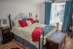 A bedroom with a wooden bed, a nightstand, a lamp, and a view of the outdoors through a window with curtains. - Runaway Rooster B&B -- Guest Cottage
 (Prince Edward)