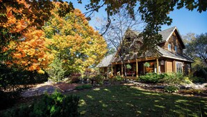 A wooden cabin with a porch, surrounded by autumn foliage and a well-maintained garden. - Runaway Rooster B&B -- Guest Cottage
 (Prince Edward)