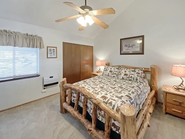 A bedroom with a rustic wooden bed frame, a ceiling fan, a window with blinds, a wardrobe, and a bedside table with a lamp. - Angel Fire Chalet park and walk (Angel Fire)