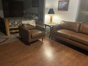 A room with a bed, two leather sofas, a small table with a lamp, and a wooden floor.
