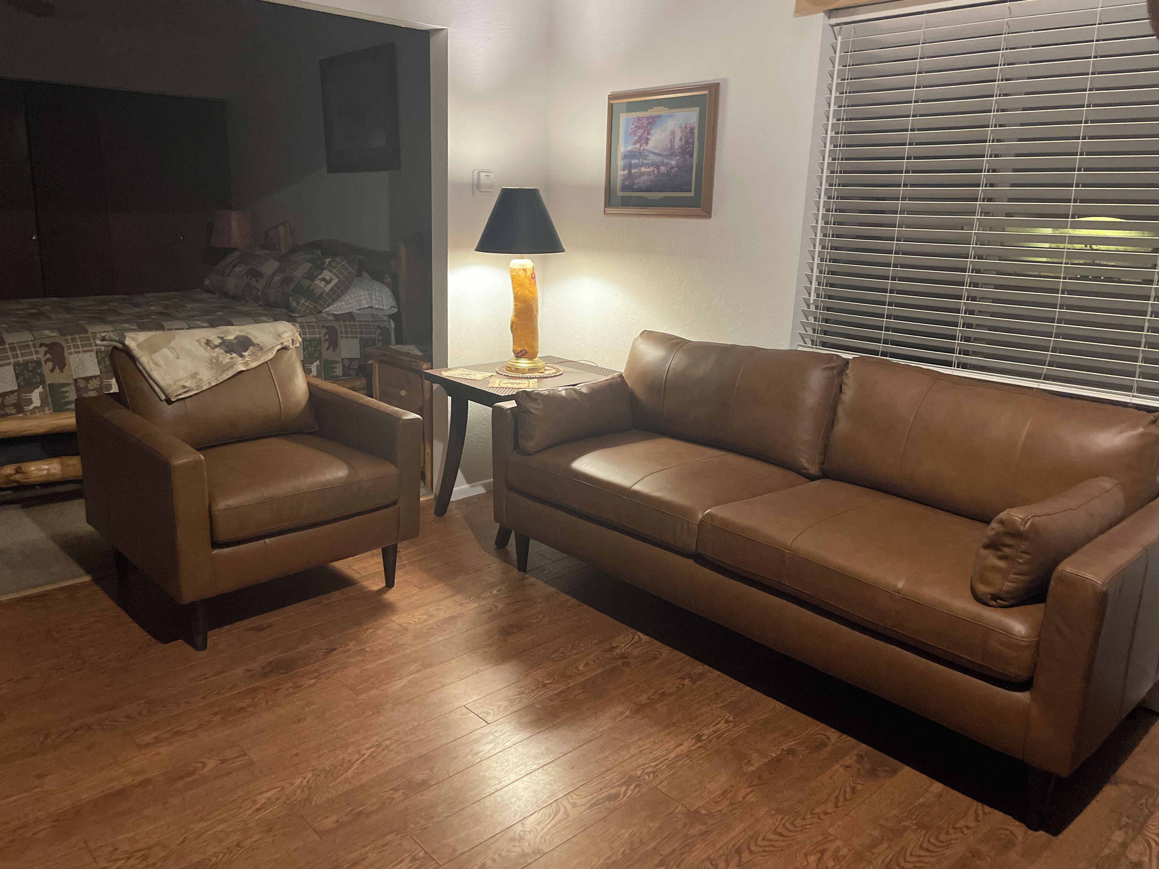 A living room with a brown leather sofa, a brown leather armchair, a small table with a lamp, and a window with blinds.