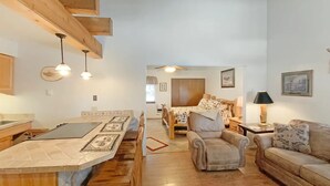 A cozy living room with a wooden dining table, a sofa, and a coffee table. There is a kitchen area in the background with a window and a door. - Angel Fire Chalet park and walk (Angel Fire)