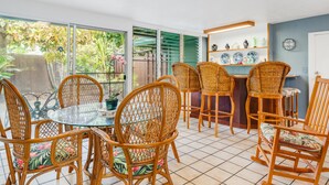A dining area with wicker chairs and a glass table, a kitchen area with a bar, and a view of greenery outside.