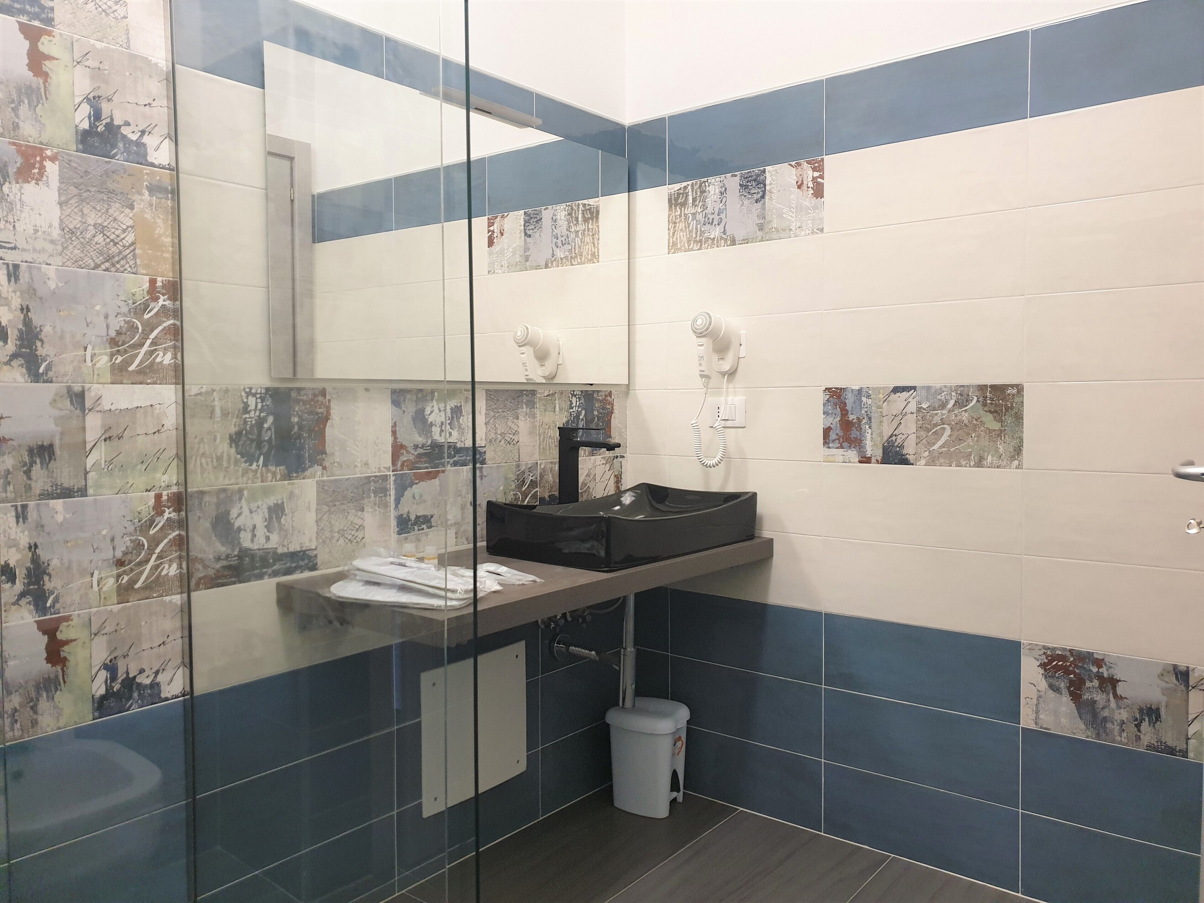 Un baño con mampara de vidrio, un lavamanos con encimera negra, un secador de pelo y un cubo de basura.