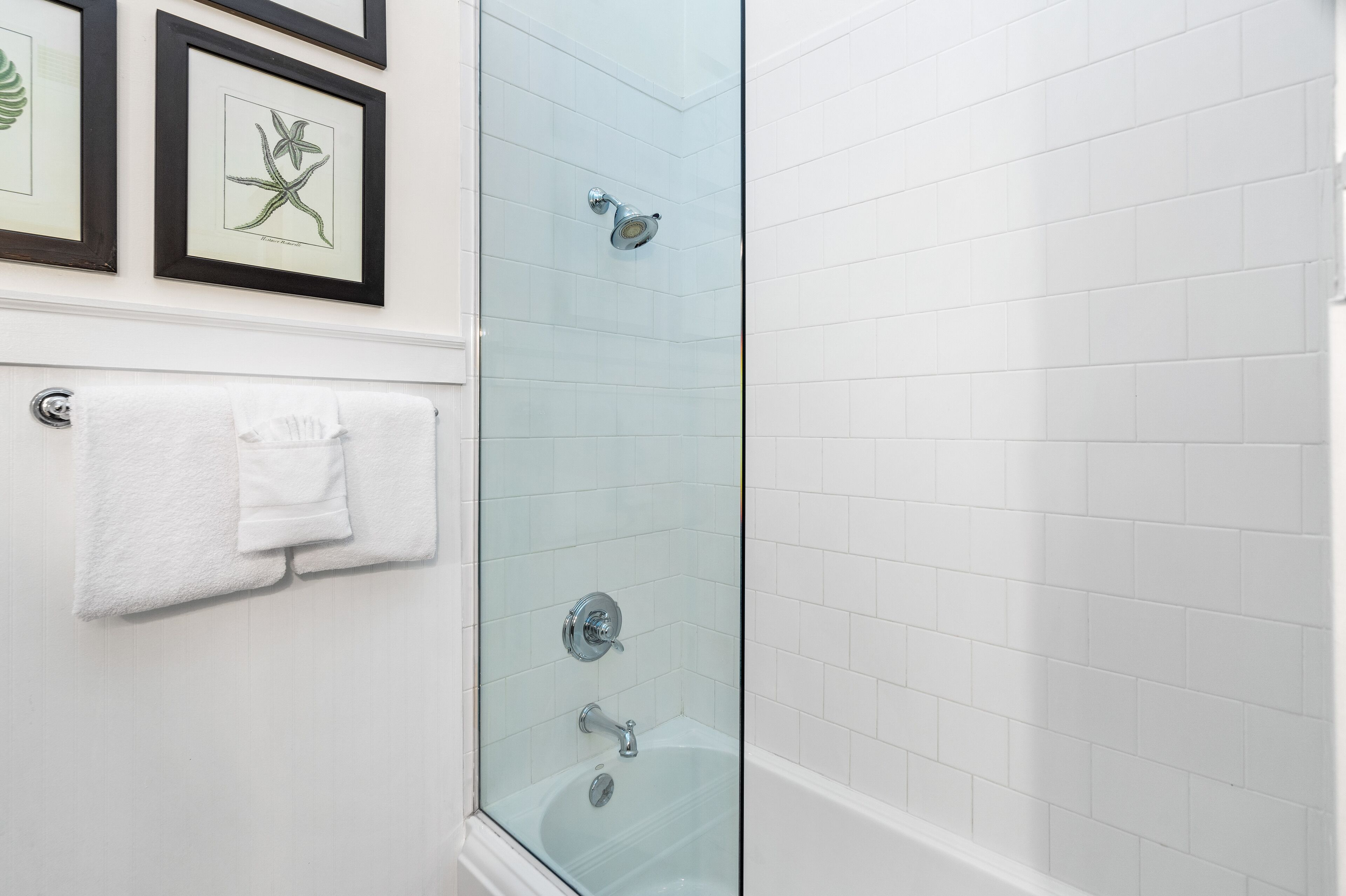 Une salle de bain dotée d’une douche à parois de verre, de murs recouverts de carreaux blancs et de reproductions botaniques encadrées.