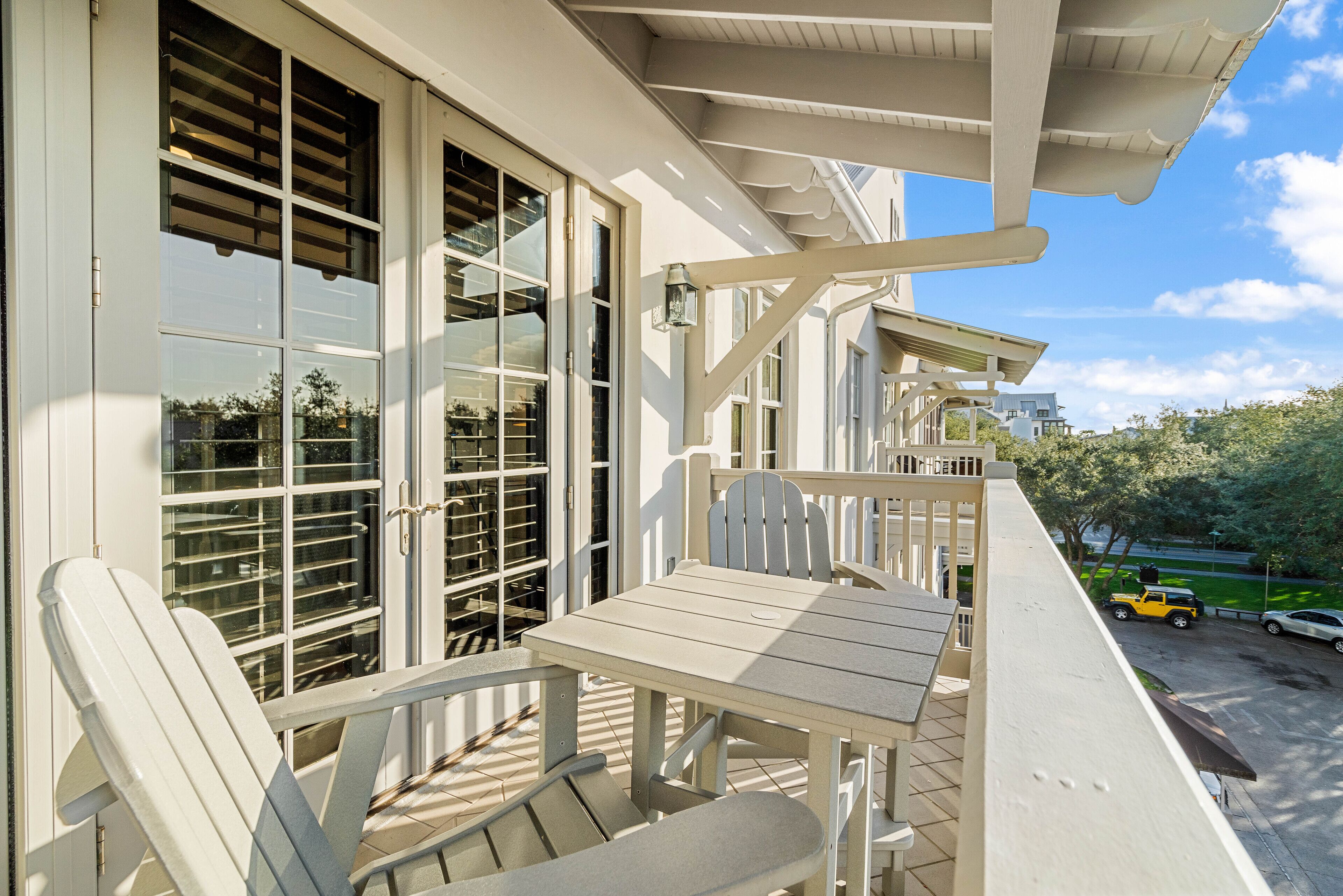 A balcony with a table and chairs, large windows with blinds, and a view of a parking area and greenery.