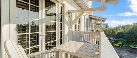 A balcony with a table and chairs, large windows with blinds, and a view of a parking area and greenery.