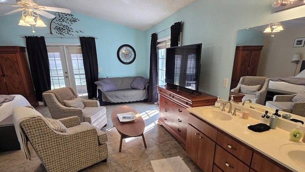 A spacious living area with a sofa, armchairs, a coffee table, a TV, and a kitchen with wooden cabinets and a sink.