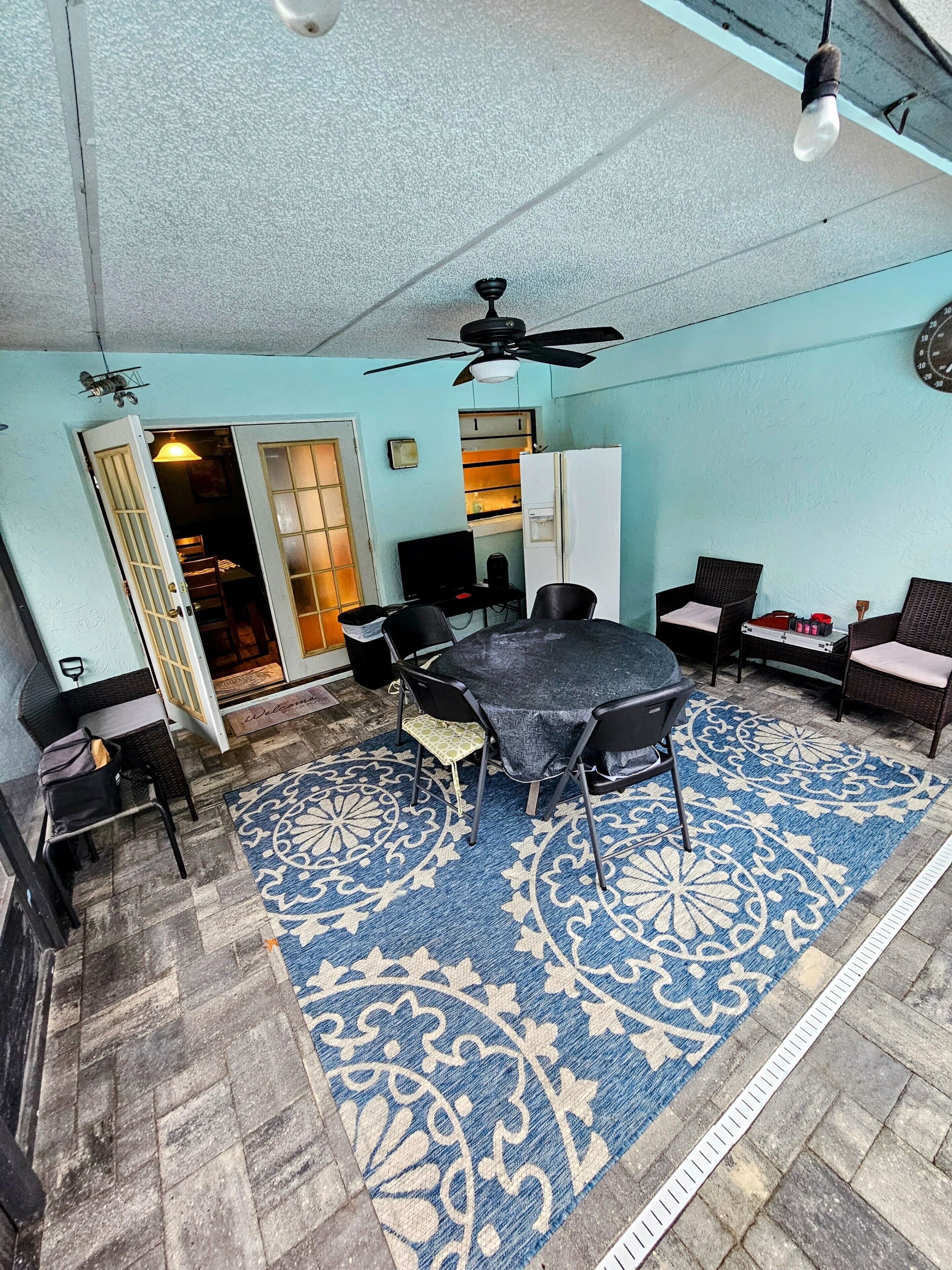 A room with a dining table, chairs, a ceiling fan, a refrigerator, and a patterned rug.