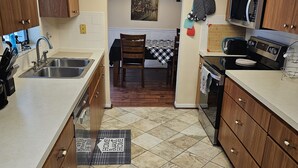 A kitchen with wooden cabinets, a stainless steel sink, a microwave, and an oven. There is a dining table with chairs and a rug in the center of the room.