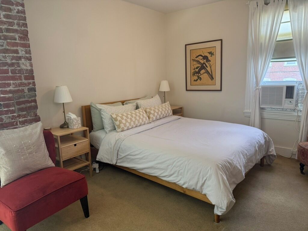 Un dormitorio con una cama, mesitas de noche, una silla roja, una lámpara y un ventanal con cortinas.
