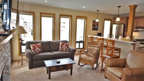 A living room with a sofa, coffee table, and patterned chairs. There is a kitchen area with a refrigerator and wooden cabinets. - Sawtooth Condo at Warm Springs Base (Ketchum)