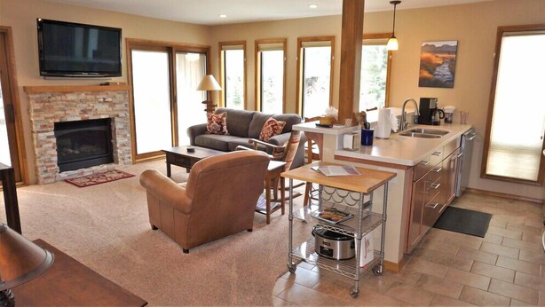 A modern living room with a fireplace, a sofa, a dining area, and a kitchen with a sink and a cart. - Sawtooth Condo at Warm Springs Base (Ketchum)