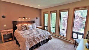 A bedroom with a large bed, a nightstand, a dresser, and a window with a view of a house and trees. - Sawtooth Condo at Warm Springs Base (Ketchum)