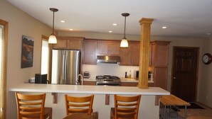 A modern kitchen with wooden cabinets, stainless steel appliances, a central island with bar stools, pendant lighting, and a clock on the wall. - Sawtooth Condo at Warm Springs Base (Ketchum)