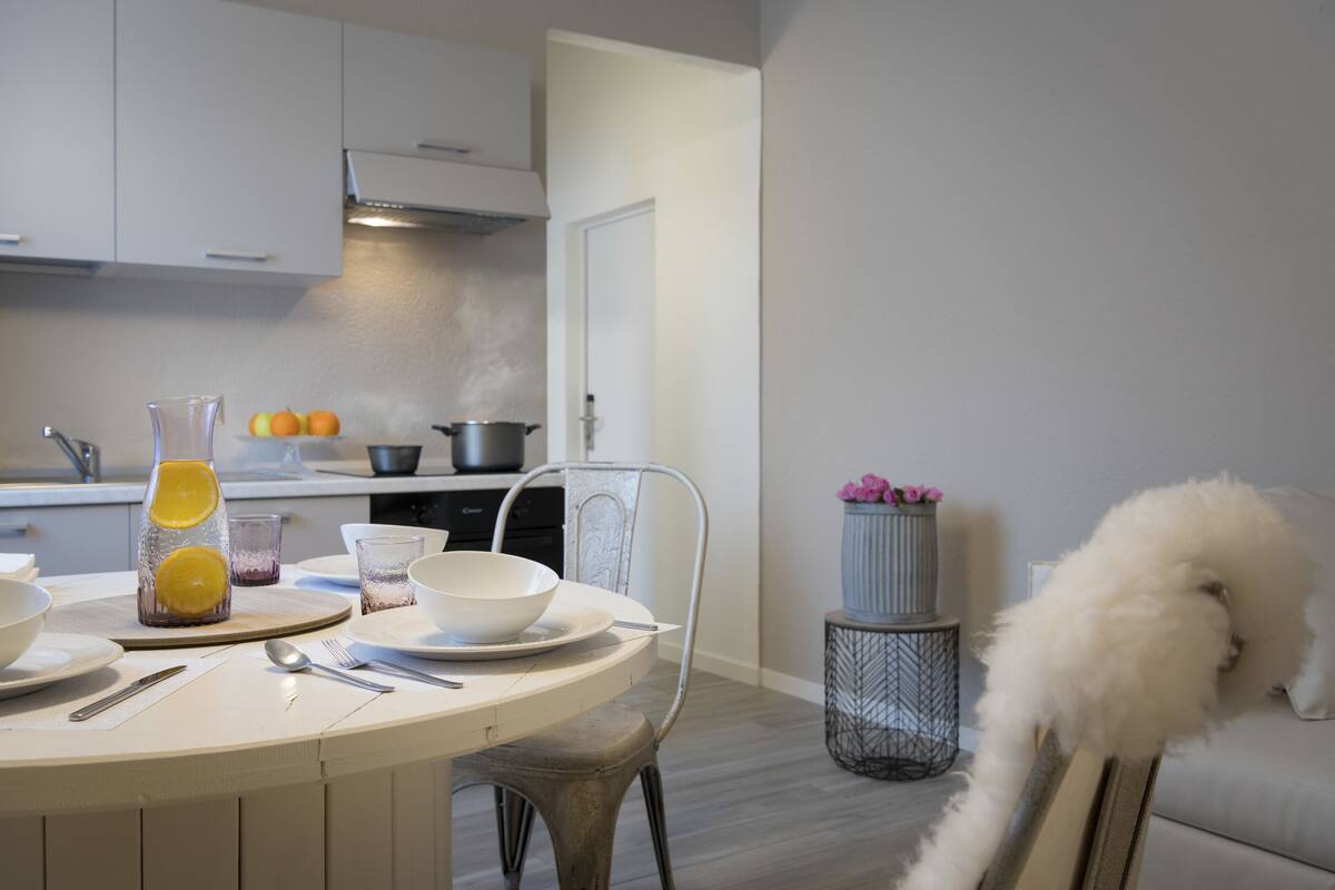 Una cocina moderna con gabinetes blancos, una mesa de comedor con platos, una jarra y un bol. Hay un perro blanco y esponjoso cerca del sofá.