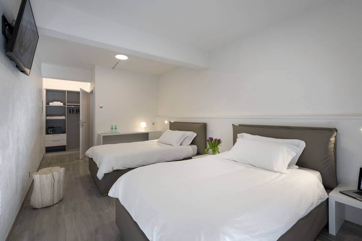 Una habitación de hotel con dos camas, un televisor montado en la pared, una mesita de noche con una lámpara y un pequeño taburete.