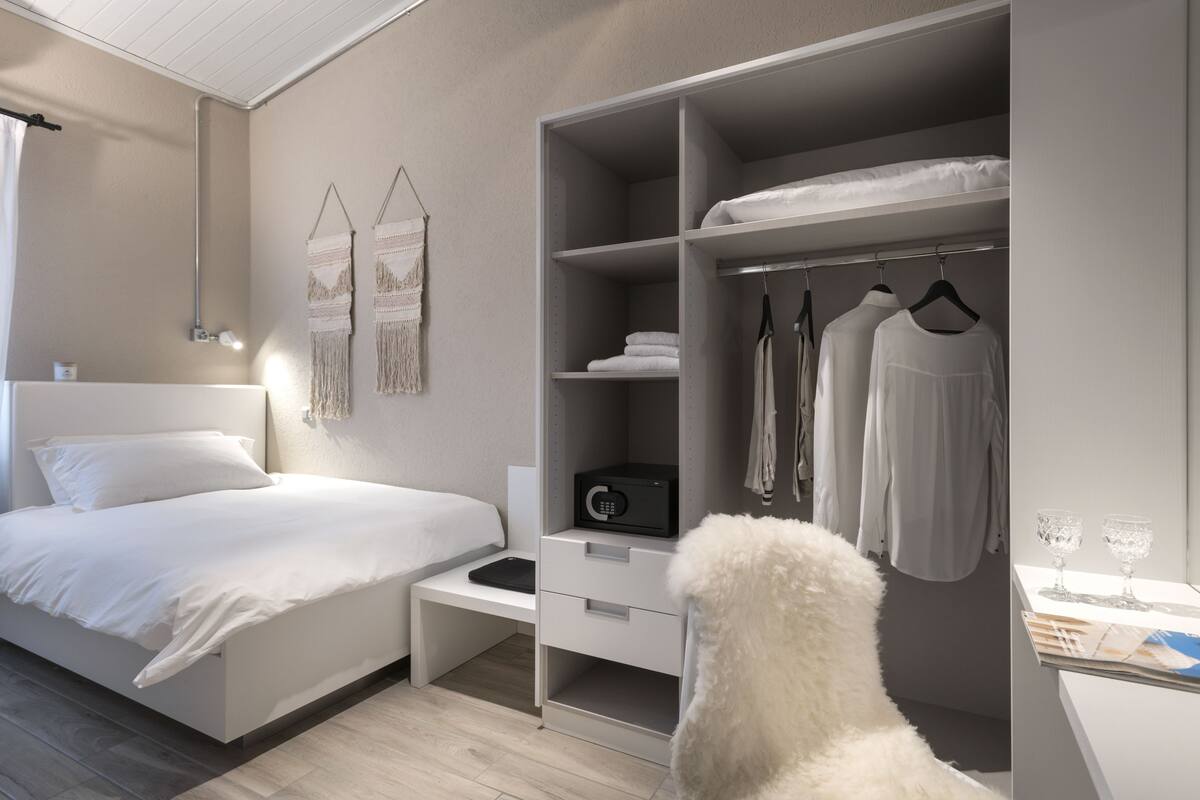 Un dormitorio con cama, mesita de noche, armario blanco y una cómoda silla blanca.