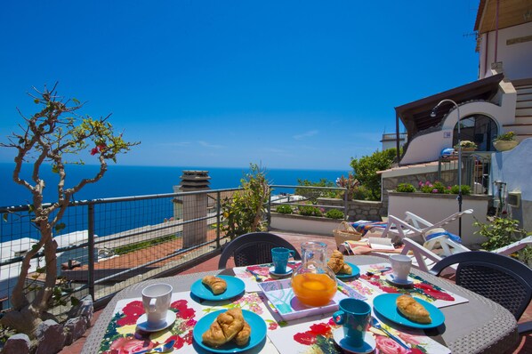 A table set with croissants, a pitcher of orange juice, and a cup, overlooking a clear blue sea.