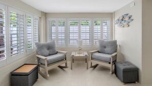 A room with two grey armchairs, a small table, a grey ottoman, and a window with shutters.