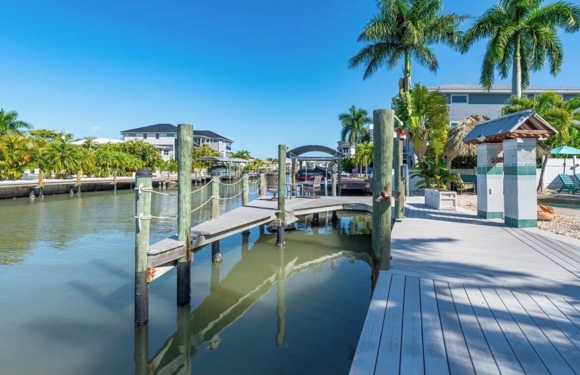A waterfront dock with a wooden walkway, palm trees, and a small pavilion.