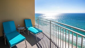 Un balcone con due poltrone blu e un tavolino, con vista su una spiaggia e un mare cristallino sotto un cielo luminoso.