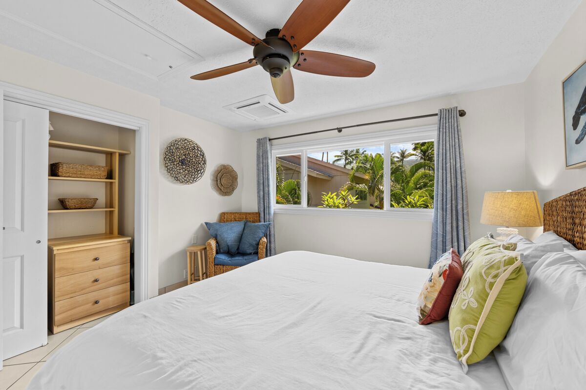Un dormitorio con cama, ventilador de techo, ventana con cortinas, una silla y mesita de noche con lámpara.