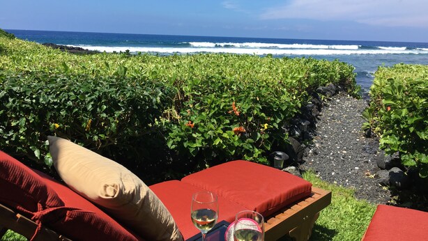 Eine Strandlounge mit roten Sitzkissen und einem Tisch mit Gläsern Weißwein, mit Blick auf den Ozean.