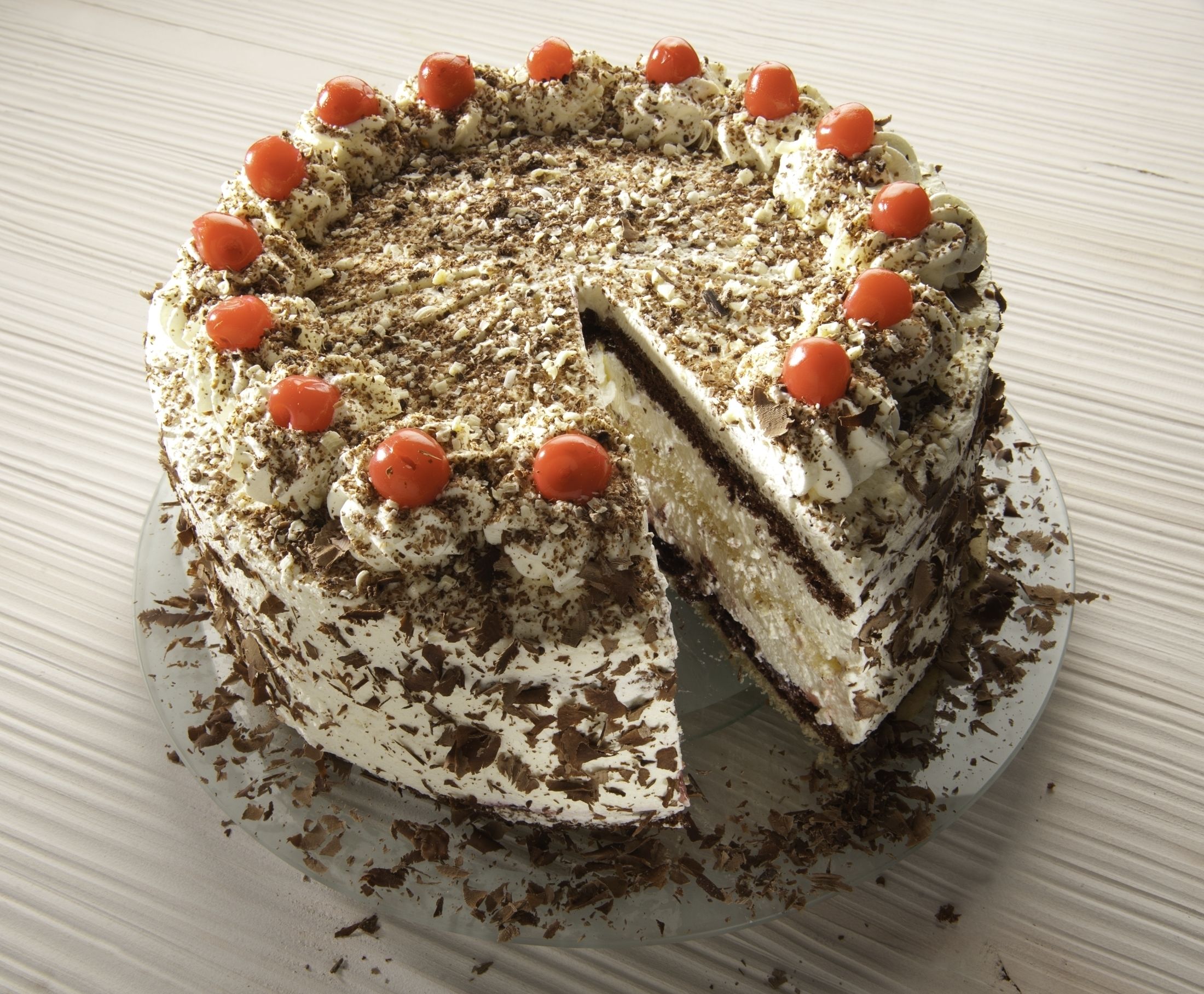 A round cake with white frosting, chocolate shavings, and red cherries on a plate.