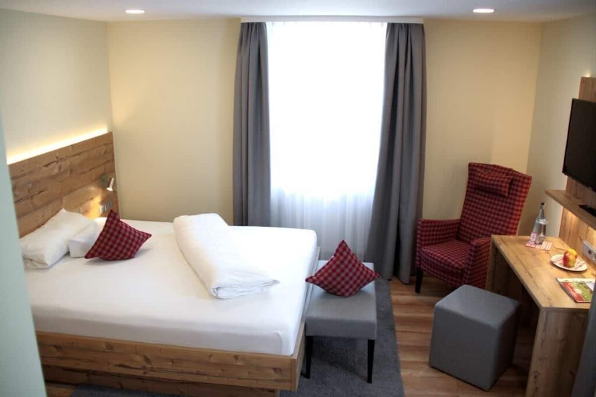 Une chambre d’hôtel avec un lit en bois, un bureau avec une chaise, une télévision et une fenêtre avec des rideaux.