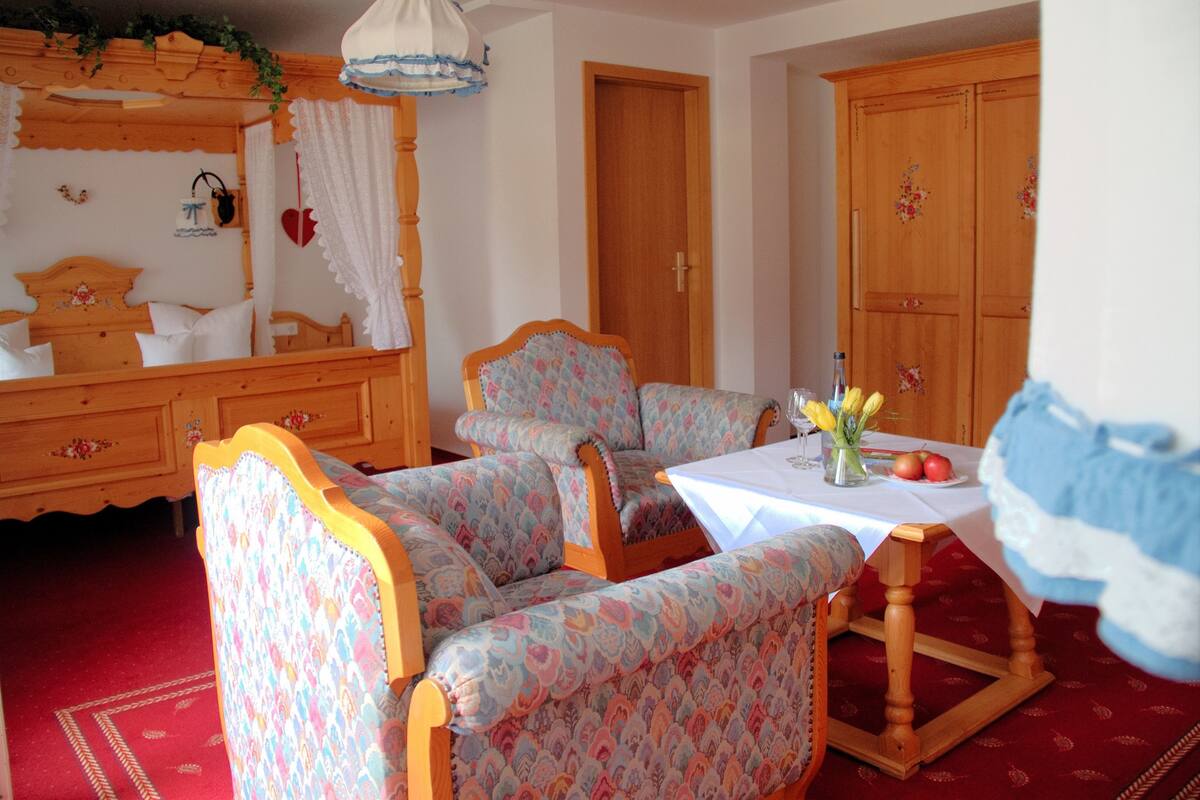 Une pièce avec un lit en bois, une chaise en bois, une table ornée d’un vase de fleurs et un tapis rouge.