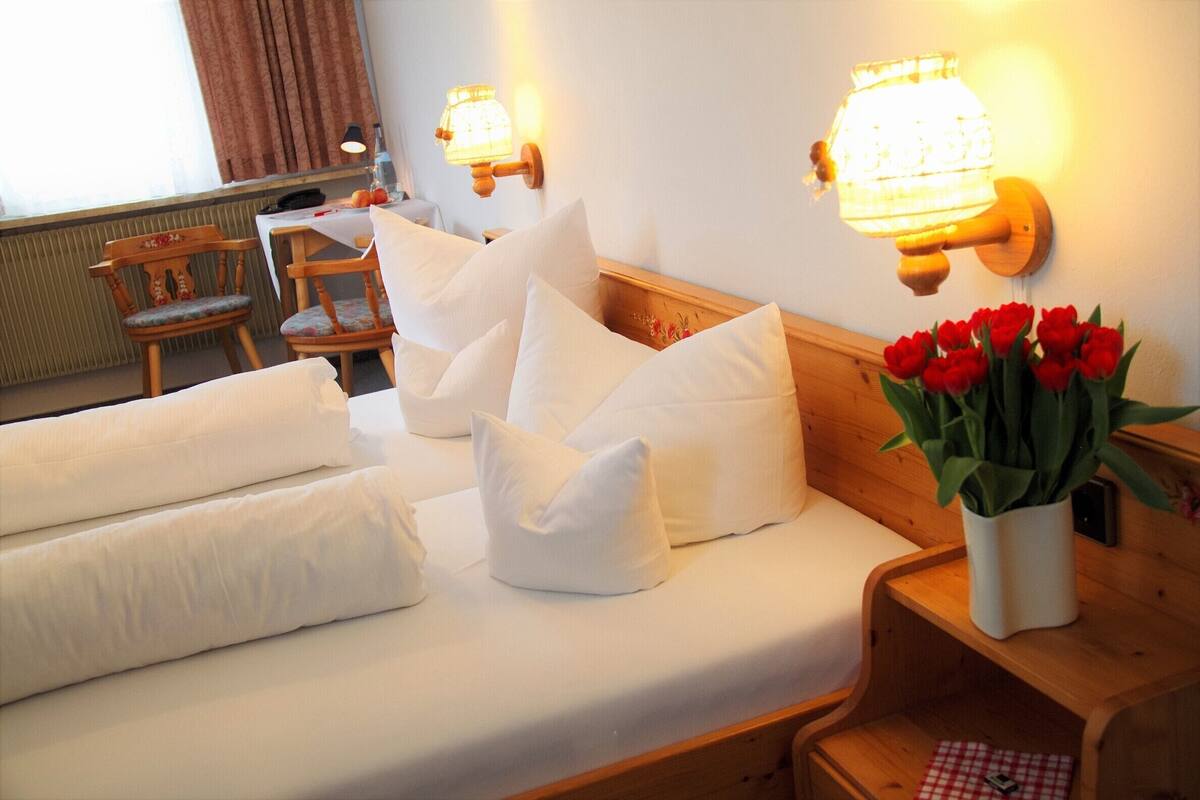Un lit bien fait, avec du linge de lit blanc et des oreillers, une table de chevet en bois ornée d’un vase rempli de fleurs rouges, et deux lampes fixées au mur.