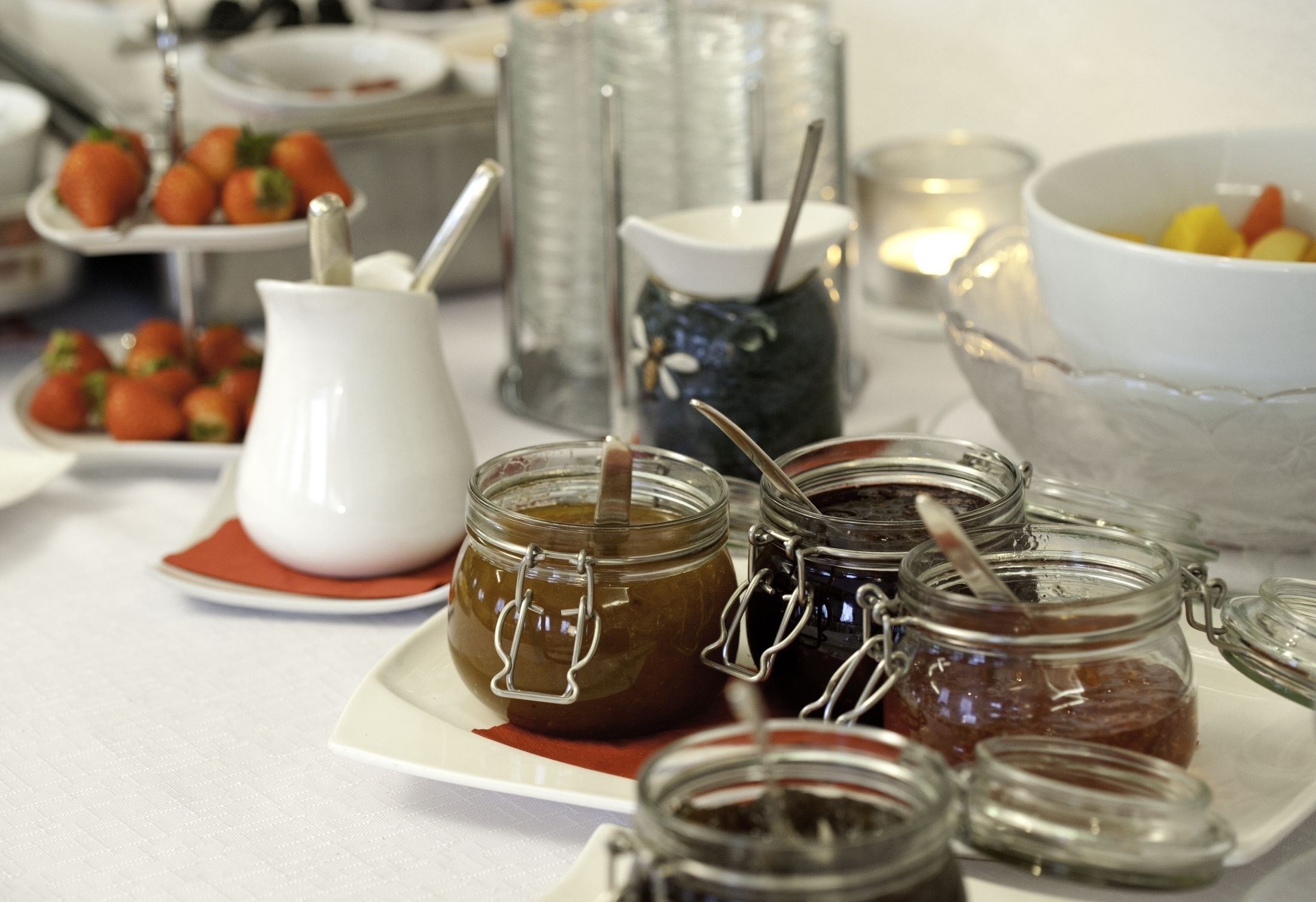 A table with various food items including jars of preserves, a pitcher, and a bowl of fruit.