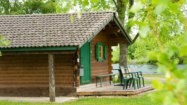 A rustic wooden cabin with a tiled roof, a green door, and a small porch with a bench and chairs. - Chalets Etape du Camping Le Bivouac (Pont-du-Navoy)