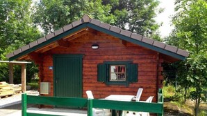 A wooden cabin with a green door and shutters, a small porch with white chairs, and a covered outdoor seating area. - Chalets Etape du Camping Le Bivouac (Pont-du-Navoy)
