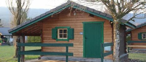Una cabaña de madera con puerta y contraventanas verdes, un porche con banca y un árbol delante.
