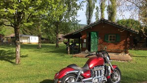 A red motorcycle parked on a dirt path in front of a rustic wooden cabin with a green door and a porch. - Chalets Etape du Camping Le Bivouac (Pont-du-Navoy)