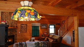 A cozy cabin interior with a fireplace, wooden walls, a staircase, and a colorful stained glass lamp.