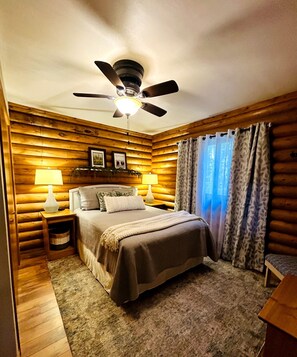 A bedroom with a bed, two bedside lamps, a ceiling fan, a wooden headboard, and a window with curtains.