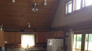 A cozy cabin interior with a wooden ceiling, a dining area with a table and chairs, a kitchen with wooden cabinets, and a large window with a view of the outdoors. - Quiet, Private and Comfortable  nestled on 8 acres of private property    (Lead)