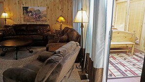 A cozy living room with a brown leather sofa, a wooden coffee table, and two floor lamps. A painting hangs on the wooden wall, and a window with blinds is visible.
