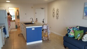 A compact living space with a kitchenette, a central island, a sofa with floral cushions, and a decorative wall piece. - Waterfront Condo Family Getaway (St Petersburg)