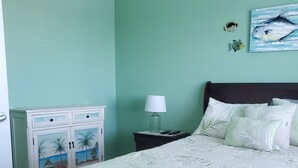 A bedroom with a bed, a nightstand with a lamp, a dresser with a beach-themed design, and a wall painting of fish. - Waterfront Condo Family Getaway (St Petersburg)