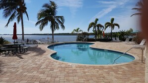 A swimming pool with a curved edge, surrounded by lounge chairs and umbrellas, with a view of the water and palm trees. - Waterfront Condo Family Getaway (St Petersburg)